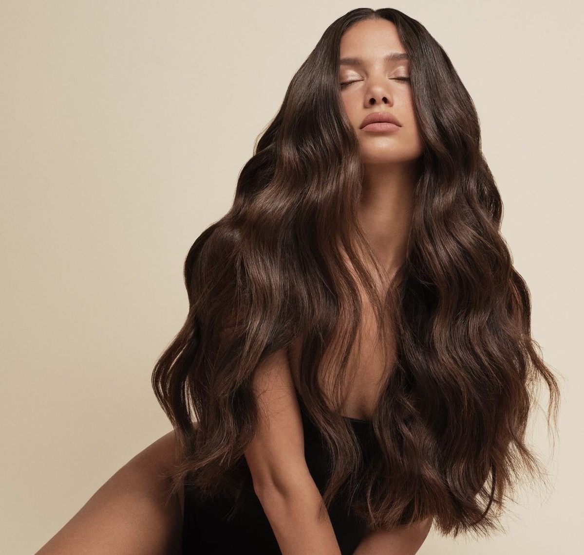 Model with long, glossy waved brunette extensions against a warm neutral backdrop